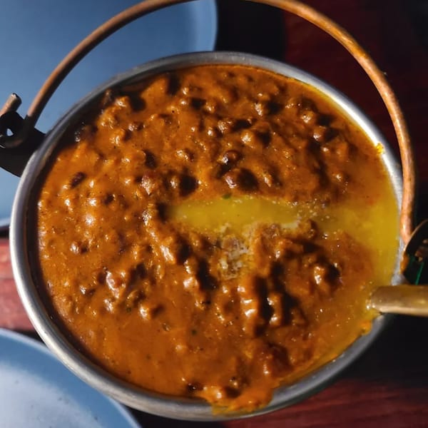 A close-up of a rich, orange Indian curry with a swirl of melted butter in a metal pot.