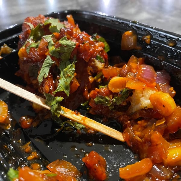 Close-up of Indian chili chicken or paneer in a black takeout container with a wooden skewer.