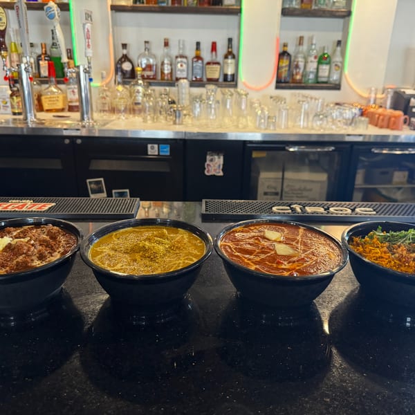 Four bowls of Indian food on a bar counter with bottles of liquor in the background.