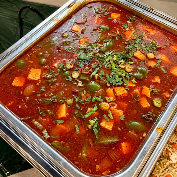 A vibrant red curry with paneer cubes, green peppers, and cilantro in a stainless steel buffet pan.