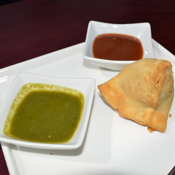 A golden-brown Indian samosa with two dipping sauces, green and red, on a white plate.
