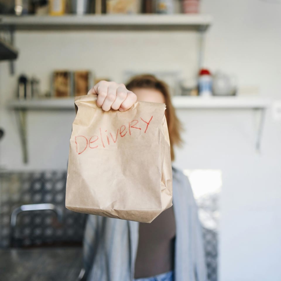 A person holds out a brown paper bag with "Delivery" written in red, in a kitchen setting.