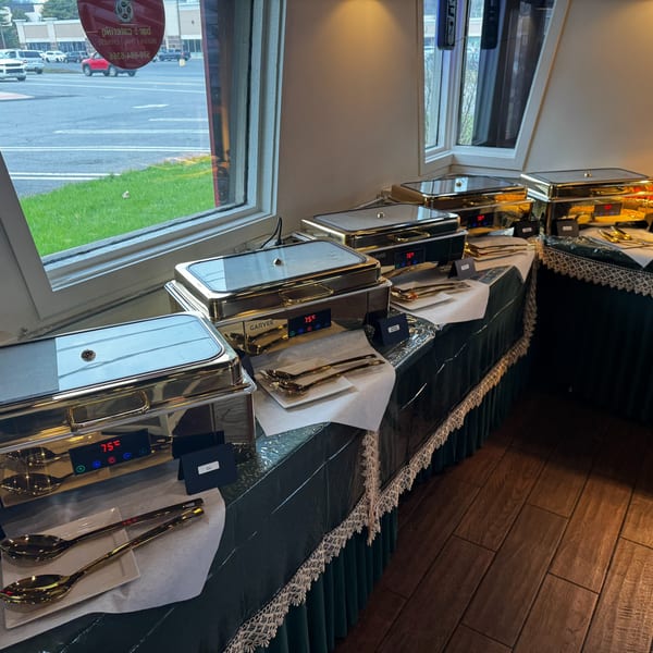 A buffet line with several silver chafing dishes and serving utensils, next to a window.
