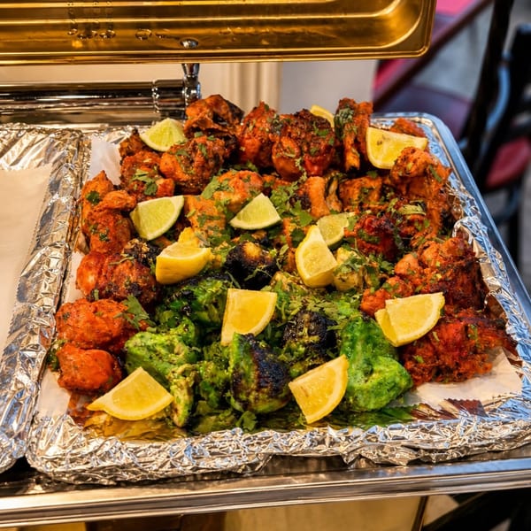 A buffet tray filled with vibrant red and green chicken tikka, garnished with fresh lime and lemon wedges.