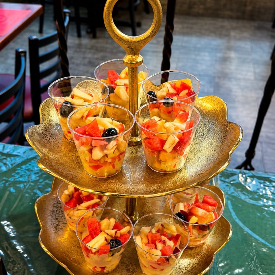 A golden tiered stand holds small cups filled with colorful diced fruit salad, including watermelon and blueberries.